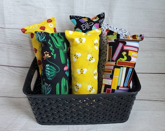 May include: A black wicker basket filled with five colorful fabric-covered pillows. The pillows have different patterns, including cacti, bees, and books.