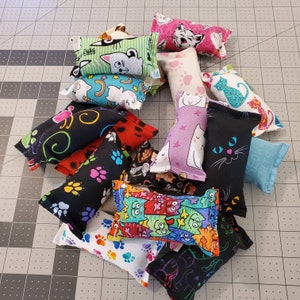 May include: A collection of cat toys in various colors and patterns. Designs include cat faces, paw prints, and abstract art. The toys are rectangular and made of fabric.