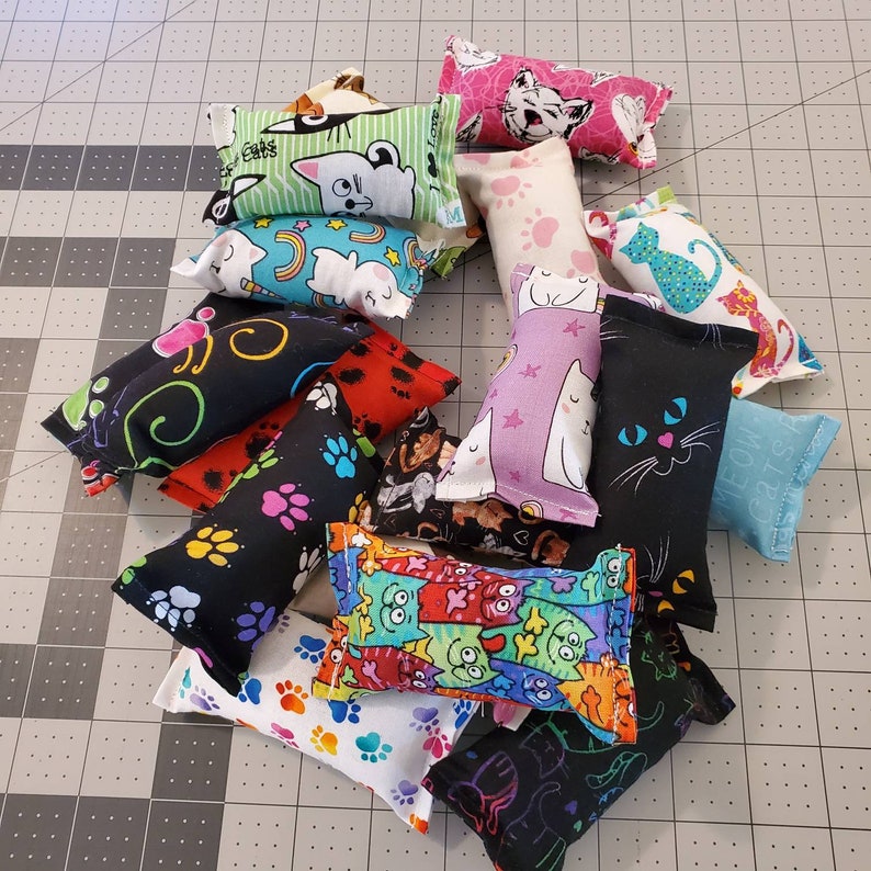 May include: A collection of cat toys in various colors and patterns. Designs include cat faces, paw prints, and abstract art. The toys are rectangular and made of fabric.