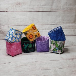 May include: Six fabric storage bins with different patterns. The bins are in a variety of colors, including pink, purple, blue, yellow, and green. The patterns include sunflowers, cacti, and daisies.