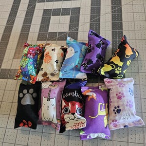May include: A collection of ten cat toys in various colors and patterns. Designs include cat faces, paw prints, and cartoon characters. The toys are made of fabric and are arranged on a gray cutting mat.