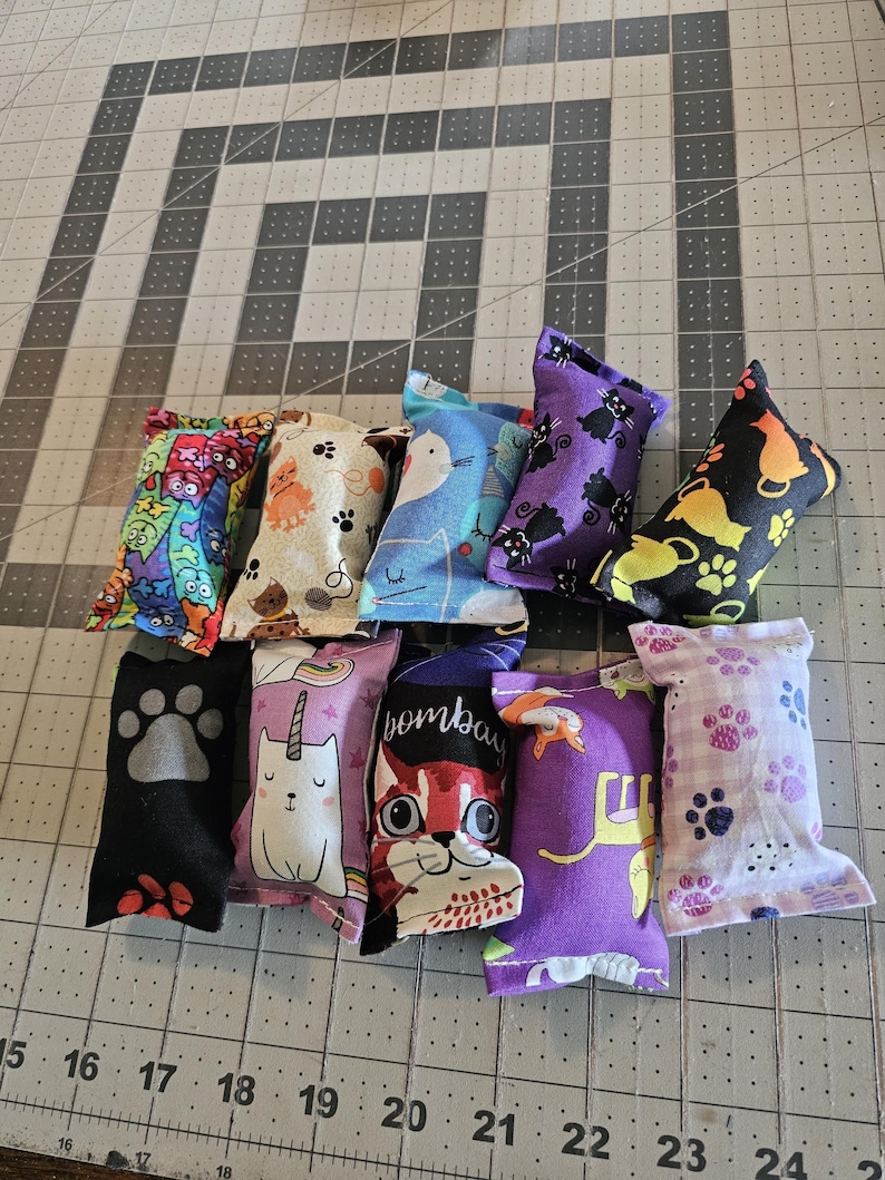May include: A collection of ten cat toys in various colors and patterns. Designs include cat faces, paw prints, and cartoon characters. The toys are made of fabric and are arranged on a gray cutting mat.