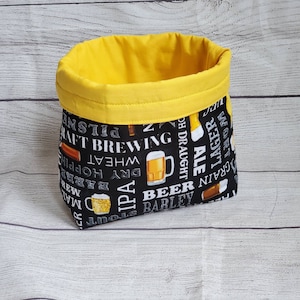 May include: A black fabric storage bin with a yellow top. The bin is printed with white text and images of beer related items, including beer bottles, hops, and barley. The text includes words like "craft brewing", "IPA", "ale", and "grain".
