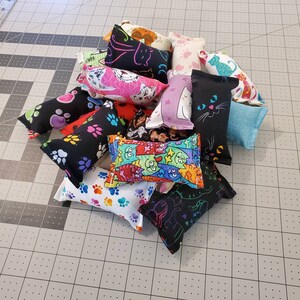 May include: A collection of cat toys in various colors and patterns. The toys are rectangular and cylindrical, with designs including paw prints, cats, and the text "Meow Cats". The toys are made of fabric and are arranged on a gray cutting mat.