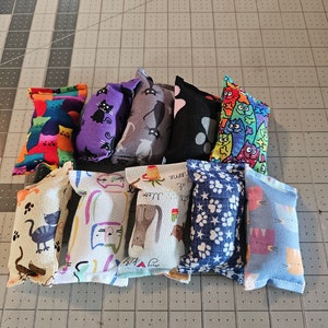 May include: A collection of catnip toys with various colorful patterns. Designs include cats, paw prints, and other whimsical illustrations. The toys are rectangular and appear to be made of fabric.