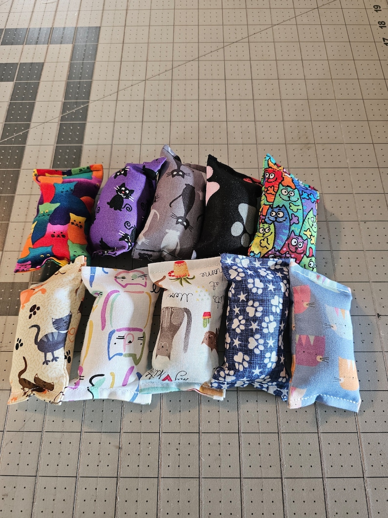 May include: A collection of catnip toys with various colorful patterns. Designs include cats, paw prints, and other whimsical illustrations. The toys are rectangular and appear to be made of fabric.