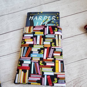 Puede incluir: Una funda de libro negra con un patr&oacute;n colorido de libros apilados y gusanos. La funda tiene un borde rojo y est&aacute; sosteniendo un libro titulado "Harper Lee" con el texto "Autor de To Kill a Mockingbird" sobre el t&iacute;tulo.
