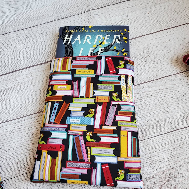 Puede incluir: Una funda de libro negra con un patr&oacute;n colorido de libros apilados y gusanos. La funda tiene un borde rojo y est&aacute; sosteniendo un libro titulado "Harper Lee" con el texto "Autor de To Kill a Mockingbird" sobre el t&iacute;tulo.