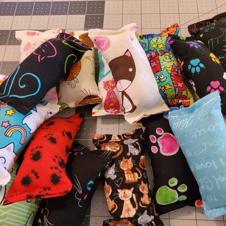May include: A collection of cat toys in various colors and patterns. The toys are rectangular and made of fabric, featuring cat-themed designs like paw prints, cats, and rainbows. Some toys are black, red, blue, and white.