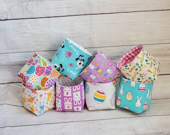 May include: Six fabric Easter baskets with different patterns. The baskets are lined up in a row. The patterns include Easter eggs, bunnies, puppies, and flowers. The baskets are all in pastel colors.
