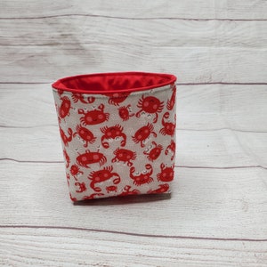 Puede incluir: Un recipiente de tela con interior rojo y exterior gris claro estampado con cangrejos rojos de dibujos animados. El recipiente tiene forma cuadrada y está sobre una superficie de madera clara.