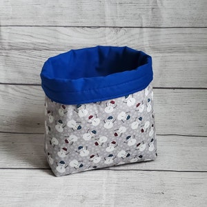 Puede incluir: Un contenedor de almacenamiento de tela gris y blanco con un forro azul. El contenedor está decorado con un patrón de muñecos de nieve.