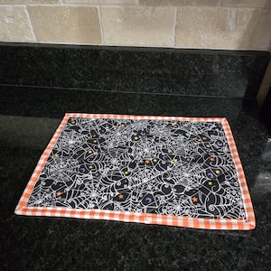 May include: Rectangular Halloween-themed placemat. The black fabric is covered in white spiderwebs, orange stars, and purple moons. The border is orange and white gingham. The placemat is on a black countertop.
