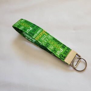Puede incluir: Un llavero verde y blanco con un anillo de llave plateado. El llavero tiene un patrón repetido de texto irlandés y tréboles. El texto incluye "Lucky", "Pot of Gold", "Shamrocks" y "Irish".
