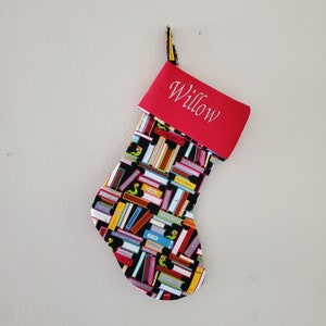 May include: Red and black Christmas stocking with a book theme. The stocking has a red cuff with the name "Willow" embroidered on it. The stocking is decorated with a pattern of colorful books and bookworms.