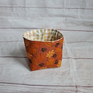 May include: A small, square-shaped fabric storage basket with a brown exterior and a gingham pattern lining. The exterior features a repeating pattern of orange and yellow leaves and brown acorns.