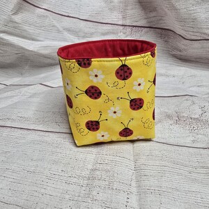 Puede incluir: Un recipiente de tela amarilla con interior rojo. El exterior está decorado con mariquitas rojas y flores blancas sobre un fondo amarillo. El recipiente tiene forma cuadrada con interior rojo.
