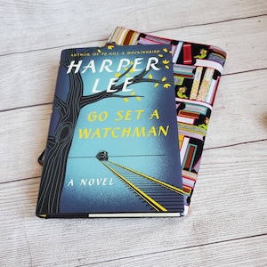 Puede incluir: Un libro de bolsillo azul y amarillo titulado "Go Set a Watchman" de Harper Lee, autora de "To Kill a Mockingbird". La portada del libro presenta un tren en una v&iacute;a de ferrocarril y un &aacute;rbol con hojas.