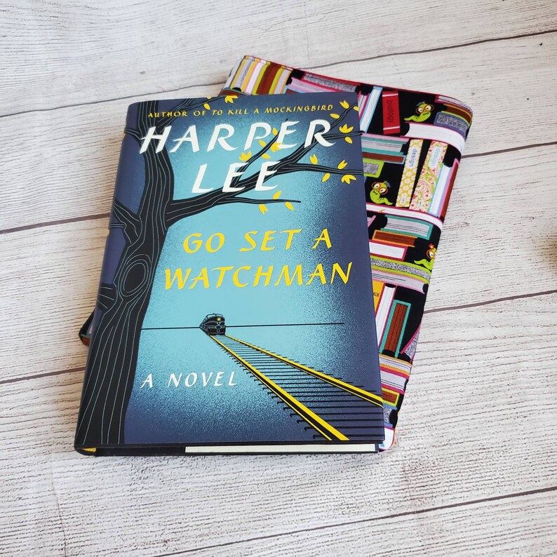 Puede incluir: Un libro de bolsillo azul y amarillo titulado "Go Set a Watchman" de Harper Lee, autora de "To Kill a Mockingbird". La portada del libro presenta un tren en una v&iacute;a de ferrocarril y un &aacute;rbol con hojas.