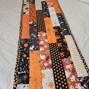 May include: A rectangular table runner featuring a patchwork design of various Halloween-themed fabrics. The patterns include skeletons, ghosts, spiders, spiderwebs, pumpkins, and black polka dots in a color palette of orange, black, and white.