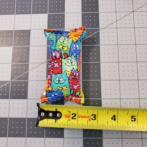 May include: A rectangular cat toy made of fabric, featuring a colorful pattern of cartoon cats in various colors. The toy measures approximately 3 inches in length.