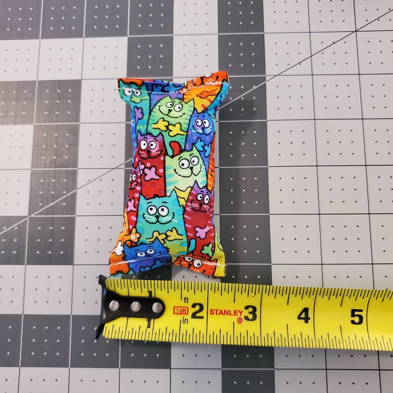 May include: A rectangular cat toy made of fabric, featuring a colorful pattern of cartoon cats in various colors. The toy measures approximately 3 inches in length.