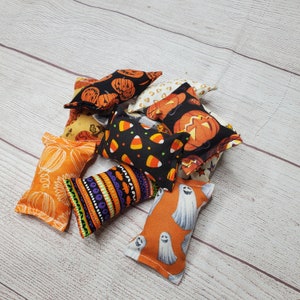 May include: A collection of nine small, handmade Halloween-themed pillows. The pillows are made of fabric with various patterns, including pumpkins, ghosts, candy corn, and stars. The pillows are all different colors, including orange, black, white, and yellow.