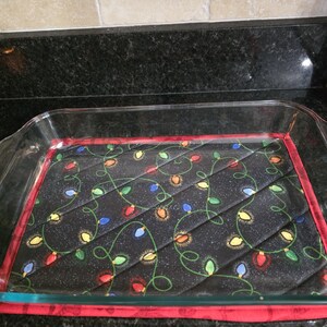 May include: A rectangular clear glass baking dish resting on a quilted fabric hot pad. The hot pad features a black background with a pattern of colorful Christmas light bulbs connected by green wires and a solid red fabric border trim.