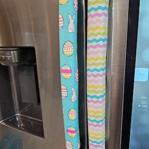 May include: Two fabric refrigerator handle covers. One is blue with white bunnies and colorful Easter eggs. The other is white with pink, yellow, and blue wavy stripes.