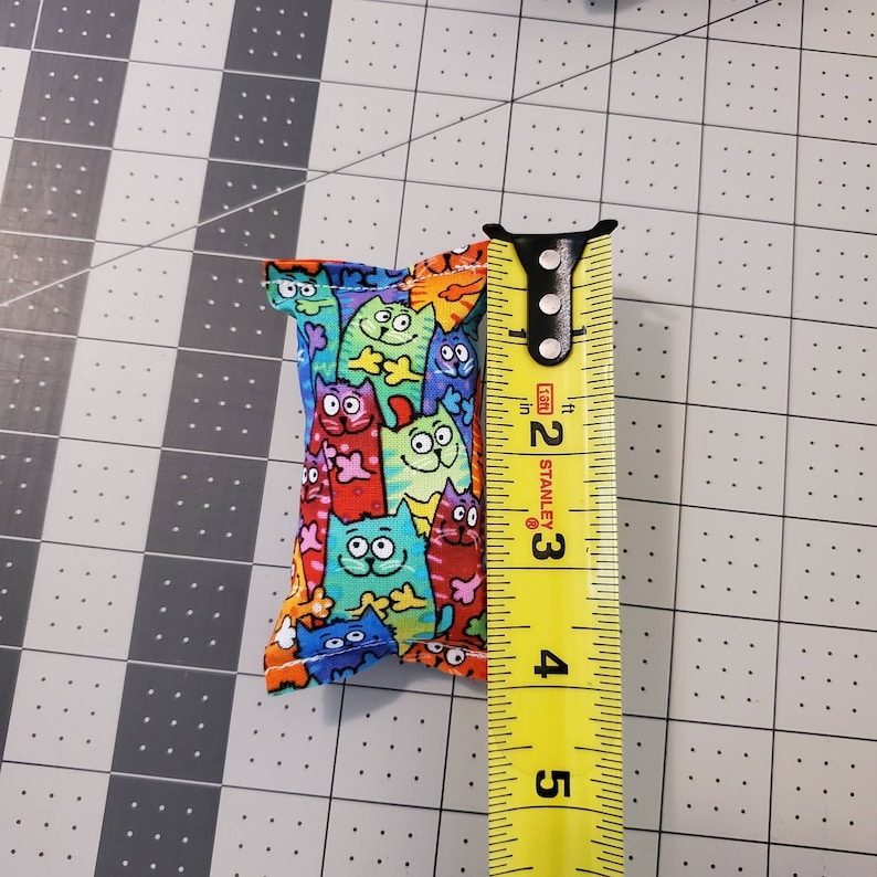 May include: A small, rectangular cat toy made of fabric, patterned with colorful cartoon cats. The toy is approximately 2 inches long. A yellow tape measure is next to the toy, showing the measurement in inches.