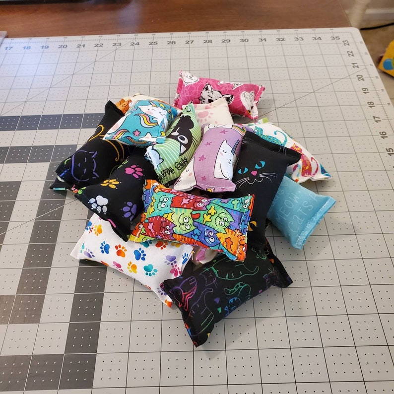 May include: A pile of cat toys in various colors and patterns. Designs include paw prints, cats, unicorns, and the text "MEOW CATS". The toys are rectangular and appear to be filled with catnip.