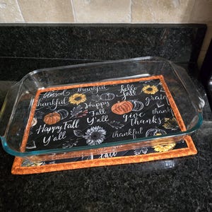 May include: A clear glass baking dish rests on a black and orange quilted trivet. The trivet features fall-themed text and graphics, including pumpkins and sunflowers, on a black background. The words "Happy Fall Y'all" are visible.