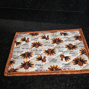 May include: A quilted rectangular trivet with a Thanksgiving theme. The trivet features a repeating pattern of cartoon turkeys in shades of brown, orange, and yellow on a white background. The words "GOBBLE 'TIL YOU WOBBLE!" are also printed on the trivet. The border is orange.
