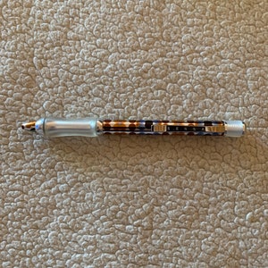 May include: A silver and brown pen with a black and white patterned barrel. The pen has a clip and a clear plastic tip. The text "SENSA" is printed on the barrel.