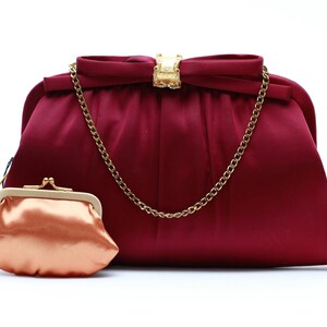 May include: A burgundy satin clutch bag with a gold chain strap and a smaller matching coin purse. The clutch features a bow detail with a gold clasp adorned with rhinestones. The coin purse is a peach colour.