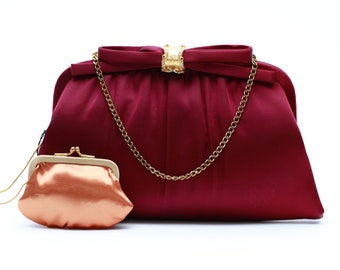Può includere: Una pochette in raso bordeaux con una catena dorata e un portamonete più piccolo abbinato. La pochette presenta un dettaglio a fiocco con una chiusura dorata ornata di strass. Il portamonete è color pesca.