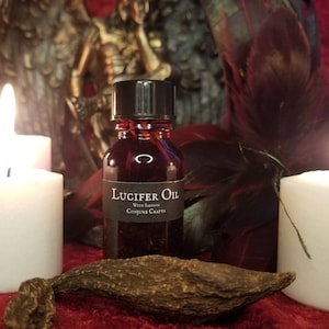 Op de afbeelding: Een kleine glazen fles "Lucifer Oil" met een zwarte dop en etiket, tegen een achtergrond van brandende witte kaarsen en donkere veren. De olie heeft een dieprode kleur. Conjure Crafts staat op het etiket.