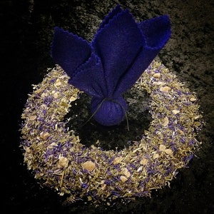 Op de afbeelding: Een cirkelvormige krans van gedroogde kruiden, paarse bloemen en kleine beige stenen omringt een diepblauwe, bloemvormige bundel. De bundel is vastgebonden met een zwart touwtje. De achtergrond is een donker, getextureerd oppervlak.