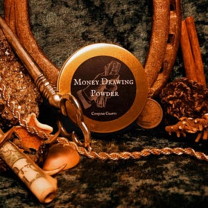 May include: An arrangement of items including a round tin labeled "Money Drawing Powder" with a hand holding money, a horseshoe, scissors, a rolled scroll, coins, and a gold chain. The items are arranged on a dark green surface.
