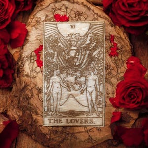 May include: A wooden tarot card featuring the Lovers card design. The card is surrounded by red roses and petals on a wooden surface. The card depicts a sun, two figures, and the text "THE LOVERS."