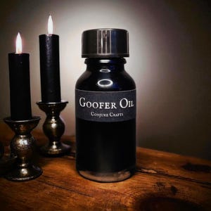 Peut inclure: Une bouteille noire d'huile de Goofer de Conjure Crafts, avec un bouchon noir, est posée sur une surface en bois. Deux bougies noires dans des supports ornés flanquent la bouteille, leurs flammes allumées. Thème mystique et rituel.