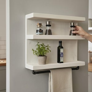May include: A white wooden kitchen shelf with three tiers, holding jars of spices, a potted plant, and a bottle of olive oil. A black towel bar is below the bottom shelf. A hand reaches for a pepper grinder.