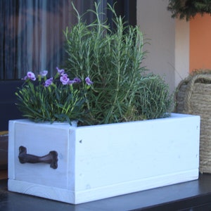 Peut inclure: Jardinière en bois blanc avec une poignée en métal. La jardinière est remplie de plantes vertes et de fleurs violettes.