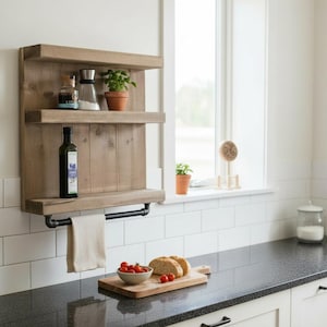May include: A rustic, wooden kitchen shelf with three tiers. The top two shelves hold spices and a potted plant. The bottom shelf has a towel rack. A bottle of olive oil and a towel hang below. The shelf is mounted on a white wall.