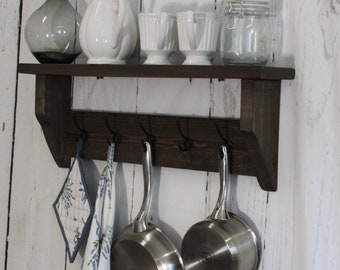May include: A dark brown wooden shelf with hooks for hanging kitchen utensils. The shelf has a white vase, a white teapot, and three white cups on it. There are two silver pots hanging from the hooks, and a white and blue tea towel is hanging on the left hook.