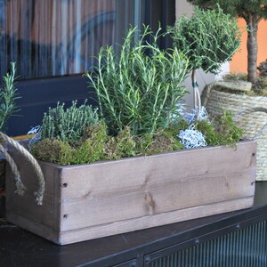 May include: A wooden planter box with a rope handle, filled with rosemary, thyme, and moss. The planter is sitting on a black metal surface.