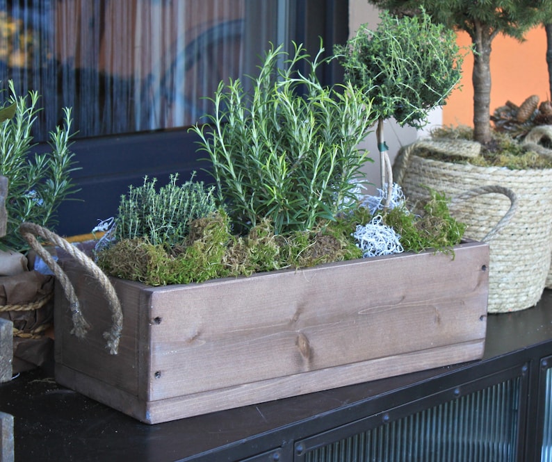 May include: A wooden planter box with a rope handle, filled with rosemary, thyme, and moss. The planter is sitting on a black metal surface.