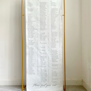 Puede incluir: Un cartel de lona blanca con un soporte de metal dorado. El cartel tiene las palabras "Nuestra gente favorita" escritas en la parte superior. El cartel enumera los nombres en orden alfabético con números junto a cada nombre.