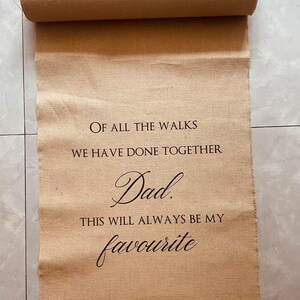 May include: A rolled-up, tan-colored fabric scroll with the text "OF ALL THE WALKS WE HAVE DONE TOGETHER Dad. THIS WILL ALWAYS BE MY favourite" in dark brown script. The scroll is unrolled, revealing the message.