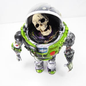 May include: A modified Buzz Lightyear action figure with a skull inside the helmet. The figure is primarily green, black, and white, with the words "Space Ranger" and "Lightyear" visible. The figure has a distressed, zombie-like appearance.